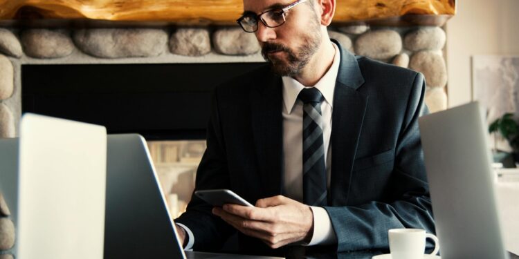 Businessman in formal attire working on a laptop and smartphone at an indoor office desk.