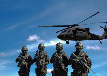 Special forces soldiers in tactical gear with helicopter overhead under a blue sky.