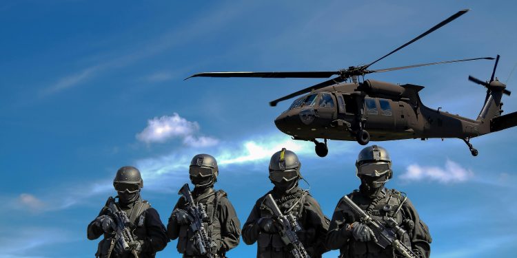 Special forces soldiers in tactical gear with helicopter overhead under a blue sky.
