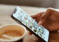 Hand browsing social media photos on a smartphone next to a cup of coffee indoors.