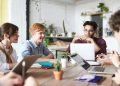 A diverse group of adults collaborating in a modern office setting with laptops and plants.