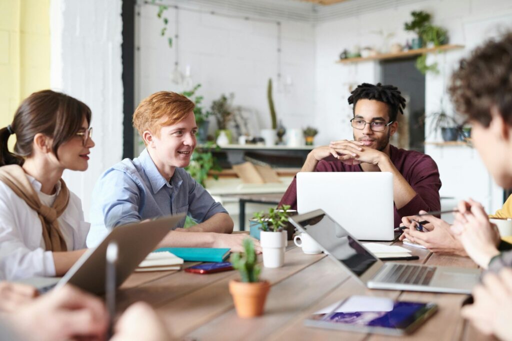 A diverse group of adults collaborating in a modern office setting with laptops and plants.