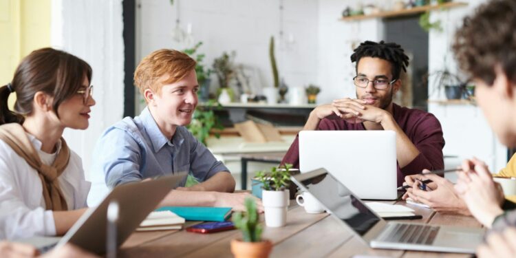 A diverse group of adults collaborating in a modern office setting with laptops and plants.