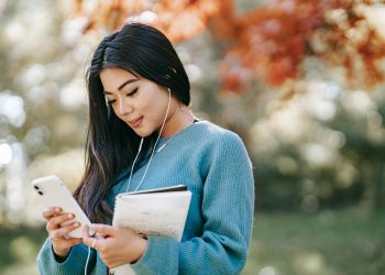Side view of young smiling Asian female student with notebook and earphones browsing mobile phone in park