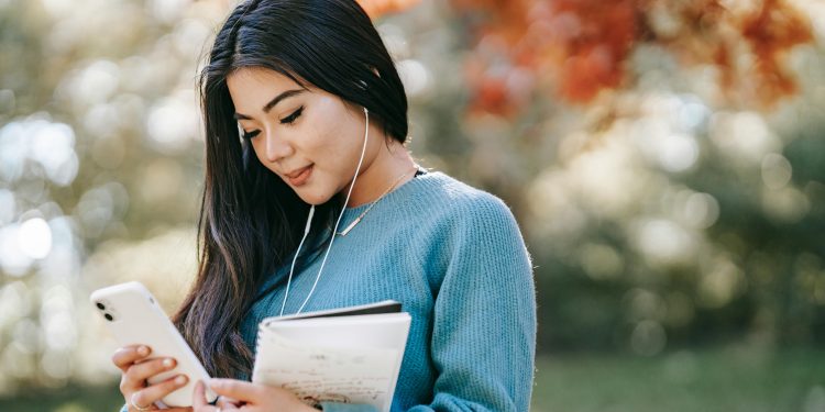 Side view of young smiling Asian female student with notebook and earphones browsing mobile phone in park