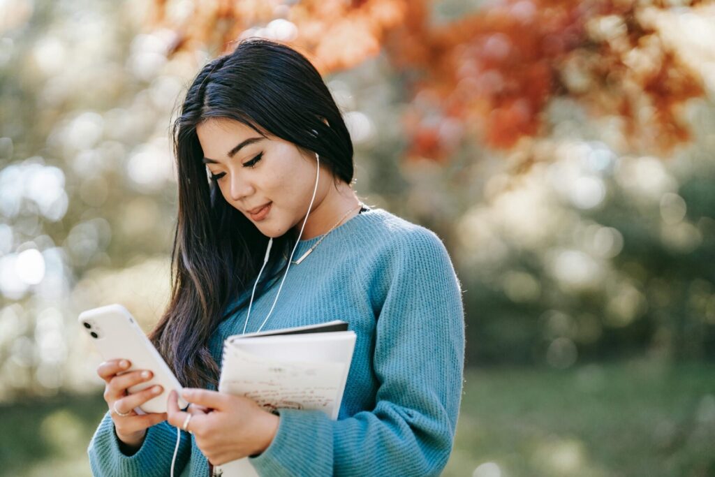 Side view of young smiling Asian female student with notebook and earphones browsing mobile phone in park