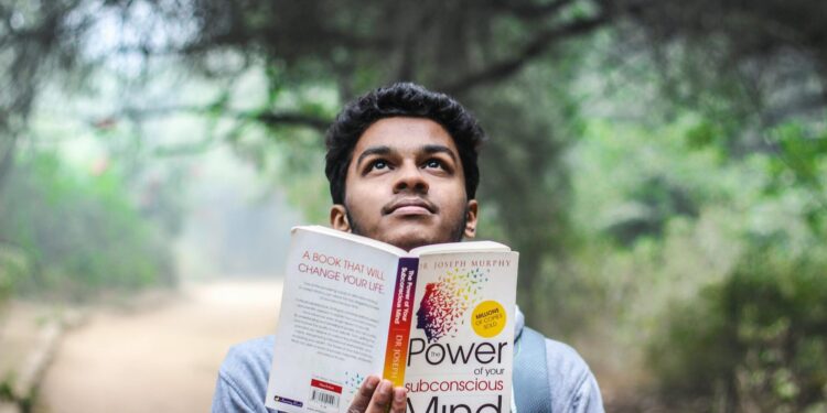 A young man relaxes while reading 'Power of Your Subconscious Mind' outdoors in a park.