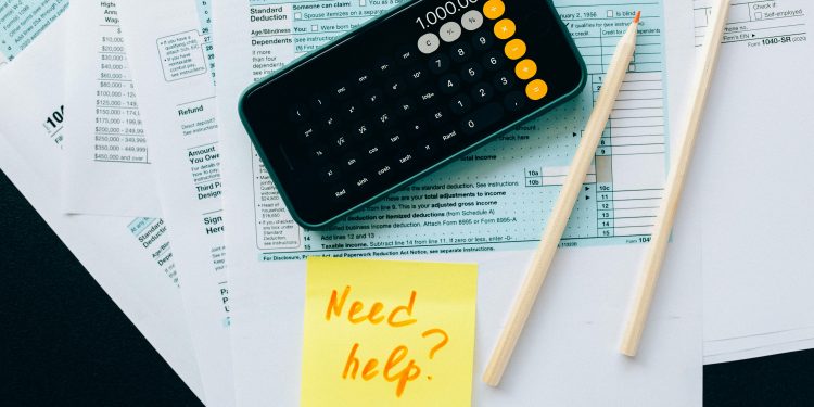 Top view of financial papers with a calculator, pencils, and a note saying 'Need help?' indicating tax or accounting assistance.