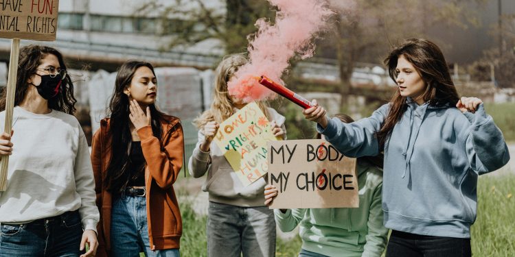 Group of women at a protest for women's rights holding signs and lighting a smoke bomb outdoors.