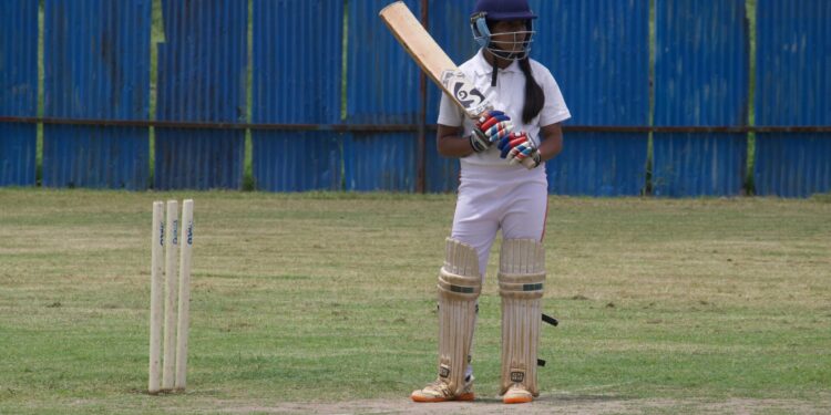 A young female cricket player poised to bat on a grassy field in Gahunje, India.