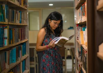 A South Asian woman with glasses reads attentively in a Delhi library, surrounded by bookshelves.