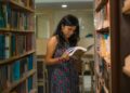 A South Asian woman with glasses reads attentively in a Delhi library, surrounded by bookshelves.