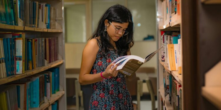 A South Asian woman with glasses reads attentively in a Delhi library, surrounded by bookshelves.