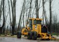 A yellow Caterpillar grader parked in a forest clearing during a cloudy day.