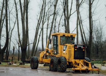 A yellow Caterpillar grader parked in a forest clearing during a cloudy day.