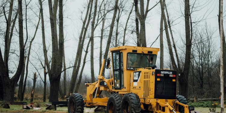 A yellow Caterpillar grader parked in a forest clearing during a cloudy day.