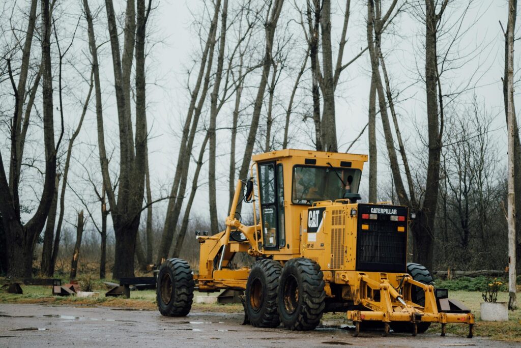 A yellow Caterpillar grader parked in a forest clearing during a cloudy day.