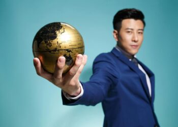 Asian businessman in blue suit confidently holding a golden globe against a blue background.