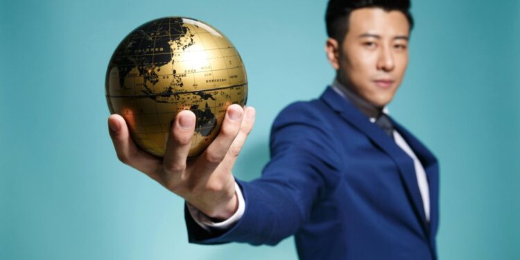 Asian businessman in blue suit confidently holding a golden globe against a blue background.