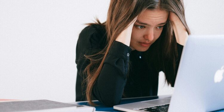 A stressed woman at a desk, looking at a laptop with a worried expression.