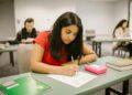 A college student concentrating while writing an exam in a classroom setting, surrounded by peers.