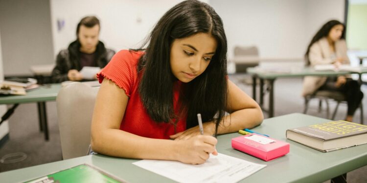 A college student concentrating while writing an exam in a classroom setting, surrounded by peers.