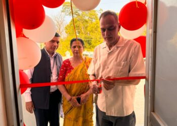 A man cuts a red ribbon at a doorway decorated with red and white balloons as two people look on.