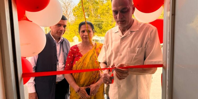 A man cuts a red ribbon at a doorway decorated with red and white balloons as two people look on.