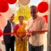 A man cuts a red ribbon at a doorway decorated with red and white balloons as two people look on.