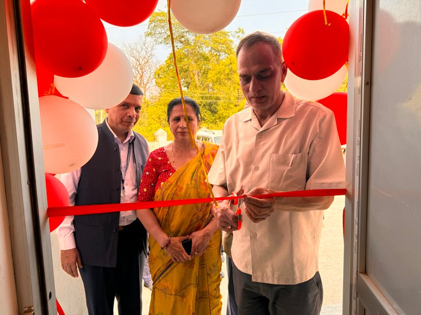 A man cuts a red ribbon at a doorway decorated with red and white balloons as two people look on.