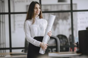 A smiling woman in a white blouse holds a large roll of paper in an office with glass partitions. Desks and office chairs are visible behind her.