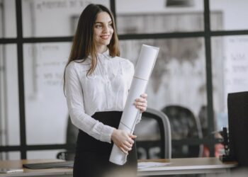 A smiling woman in a white blouse holds a large roll of paper in an office with glass partitions. Desks and office chairs are visible behind her.