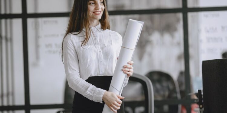 A smiling woman in a white blouse holds a large roll of paper in an office with glass partitions. Desks and office chairs are visible behind her.