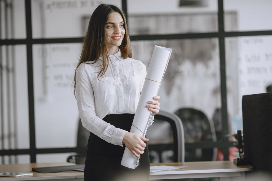 A smiling woman in a white blouse holds a large roll of paper in an office with glass partitions. Desks and office chairs are visible behind her.