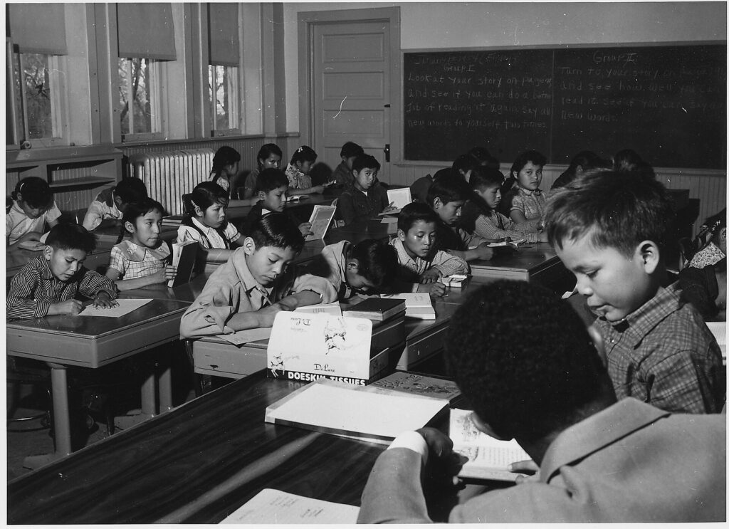 Stock photo of [One student getting assistance from teacher while other students are writing in day school.], used to illustrate this story on James Talarico’s Teaching Record Offers a Limited Political Blueprint.