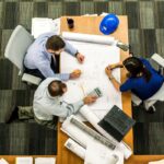 Architects and colleagues reviewing blueprints around a table, illustrating a rethink of project management.