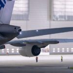 A large commercial airplane sits in an illuminated hangar with maintenance staff, illustrating airport operations ahead of the Navi Mumbai flight plan deadline.