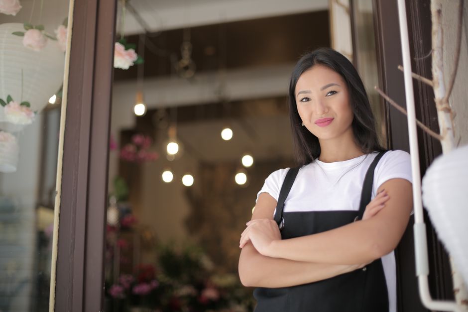 A young Asian woman stands with crossed arms at a floral shop entrance, illustrating a business loan article about cash flow decisions.