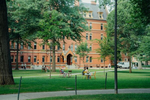 Stock photo of students relax on a lush lawn in front of a historic university building, surrounded, used to illustrate this story on Harvard’s Class of 2030 Is 43.2% Asian American and 9.3% Black.