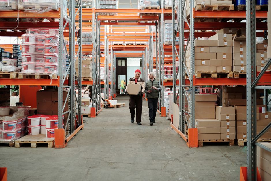 Stock photo of two workers handle a package in a spacious warehouse surrounded by shelves stocked with, used to illustrate this story on Poundland Owner Faces Administration as Reynolds Seeks Facts.