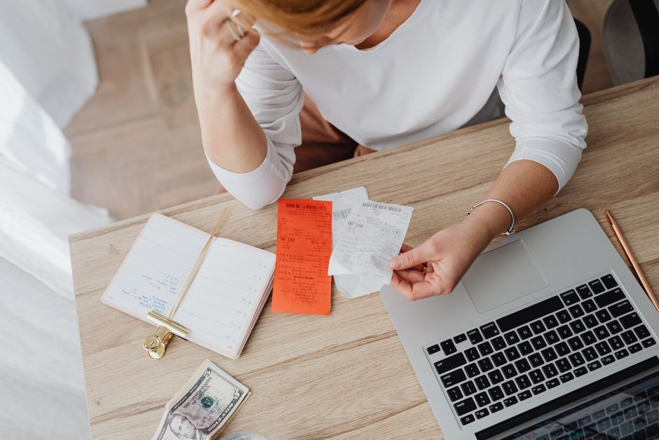 A woman reviews receipts and plans a budget with a laptop and notebook at home, illustrating household financial management amid budget changes.