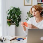 A woman reviews bills at a home desk with a laptop and plants, illustrating budget changes affecting energy bills and fuel costs.