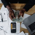 Stock photo of person counting US dollars, using a calculator and laptop, with financial documents on a, used to illustrate this story on How to Set a Salary Counter and Weigh a Job Move.