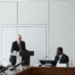 A diverse group attends a formal business meeting with a speaker at a podium, illustrating education leaders discussing early university succession.