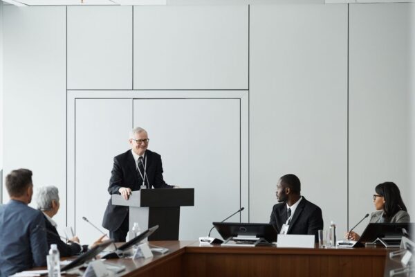 A diverse group attends a formal business meeting with a speaker at a podium, illustrating education leaders discussing early university succession.