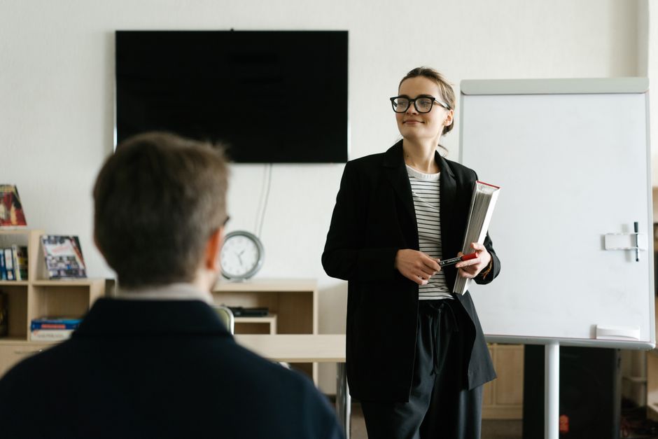 A female teacher in a black blazer presents to a student in a classroom, illustrating colleges preparing graduates with work portfolios.