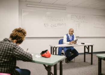 A proctor watches students taking tests in a classroom, illustrating the AP Sports Quota Teacher Recruitment exam.