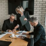Stock photo of three mature professionals in a business meeting discussing and signing documents in an office, used to illustrate this story on TVS Motor Approves Consumer Finance Demerger.