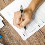 A hand writing on a W-9 tax form with a ballpoint pen on a wooden desk, illustrating small business registration paperwork.