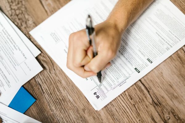 A hand writing on a W-9 tax form with a ballpoint pen on a wooden desk, illustrating small business registration paperwork.